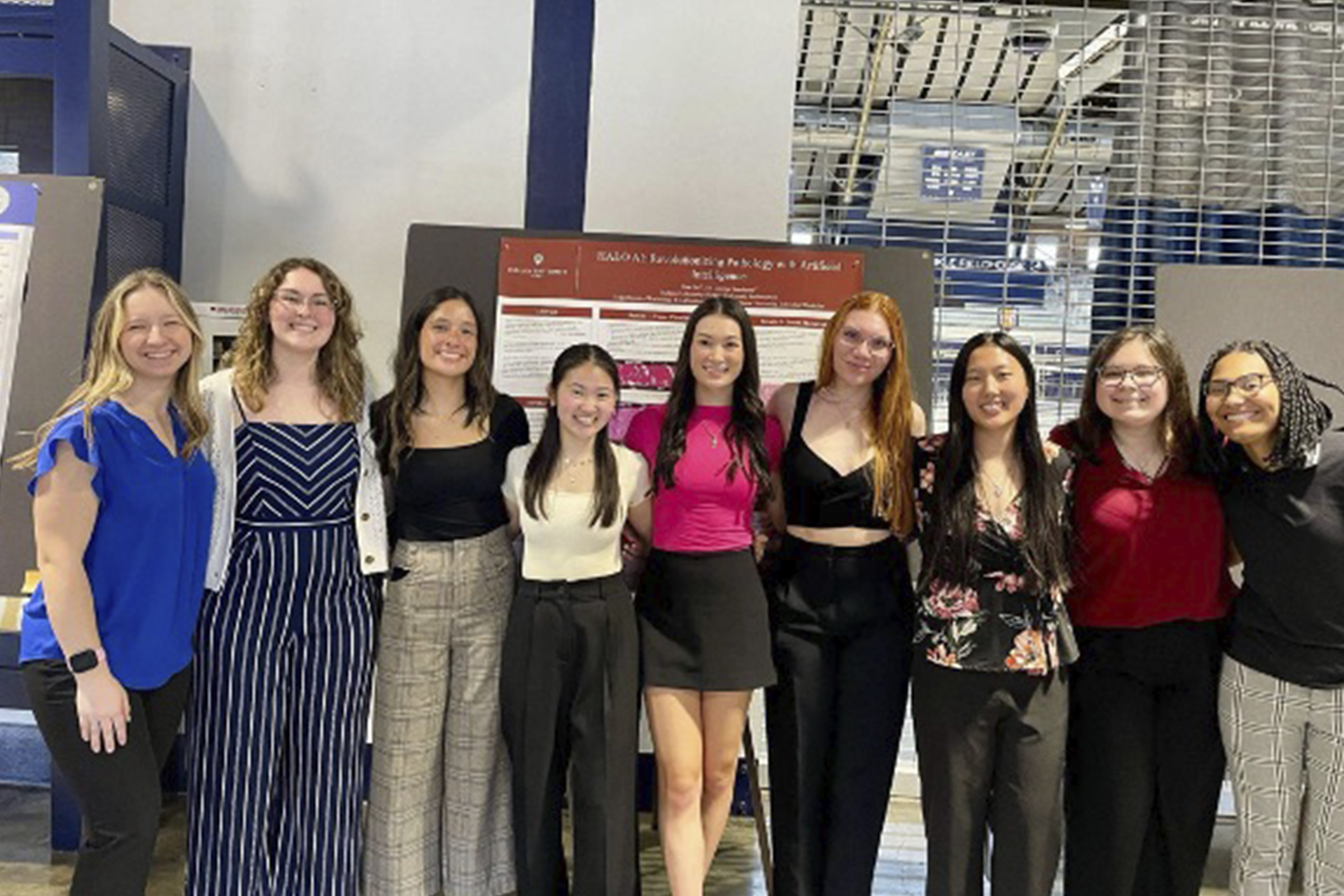 A group of nine young professionals or students standing together in a conference setting, smiling in front of a research poster presentation. They are dressed in business casual attire, with posters and industrial railings in the background.