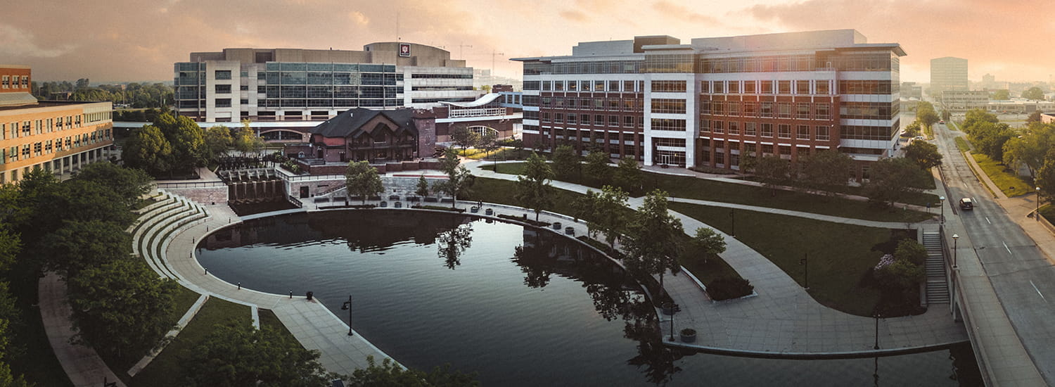 Aerial view of Indiana University campus buildings surrounding a peaceful, circular pond with a stepped amphitheater along its edge. Modern buildings with large glass windows are bathed in the warm glow of sunrise or sunset, creating a tranquil and inviting atmosphere. Trees and landscaped pathways enhance the scenic, open-campus layout.