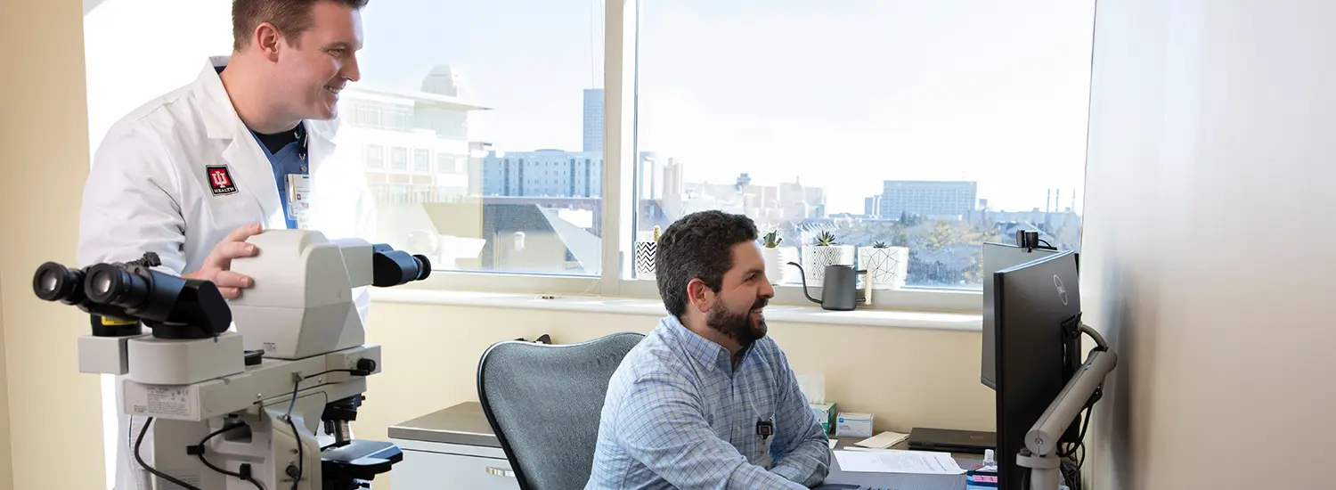 A doctor in a white lab coat stands beside a seated colleague, both smiling as they review information on a computer screen. A microscope is positioned nearby, and large windows provide natural light with a view of city buildings.