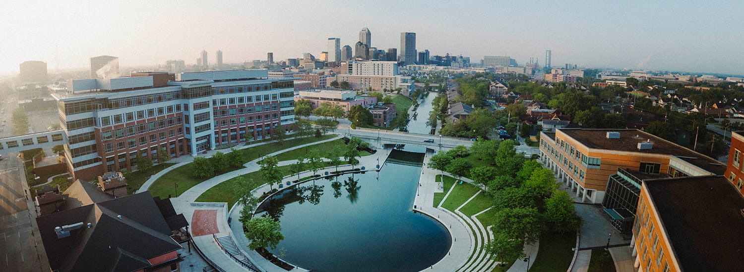Aerial view of a university campus with a circular water feature surrounded by green lawns and pathways in the foreground. Modern campus buildings line the area, and a scenic canal runs through the center, leading towards the downtown skyline in the background. The scene is calm, with a soft morning or evening light casting a glow over the cityscape.