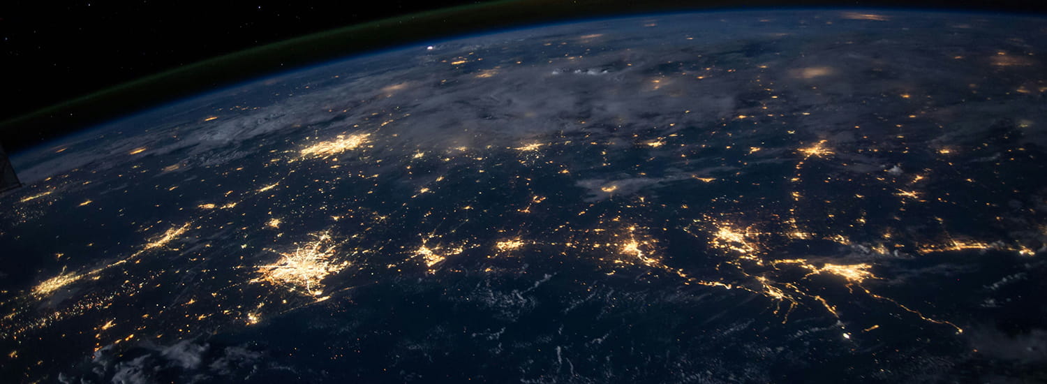 A view of Earth from space at night, showcasing illuminated cities and networks of lights across the planet's surface. The curvature of the Earth is visible, with a faint green glow of the atmosphere along the horizon. Clouds partially cover some areas, and the vastness of space extends beyond.