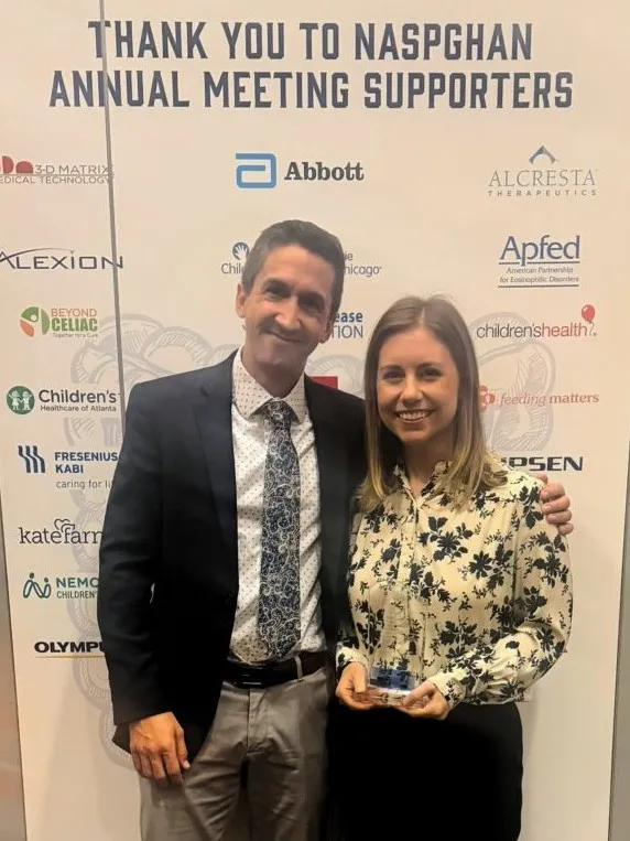 Two people stand in front of a conference banner backdrop. One is holding an award.