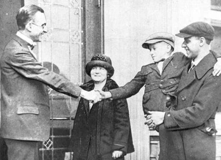 In an old black and white photo, a man holds a small boy in his arms. The boy is reaching out to shake the hand of James Whitcomb Riley. A woman looks on in the background.