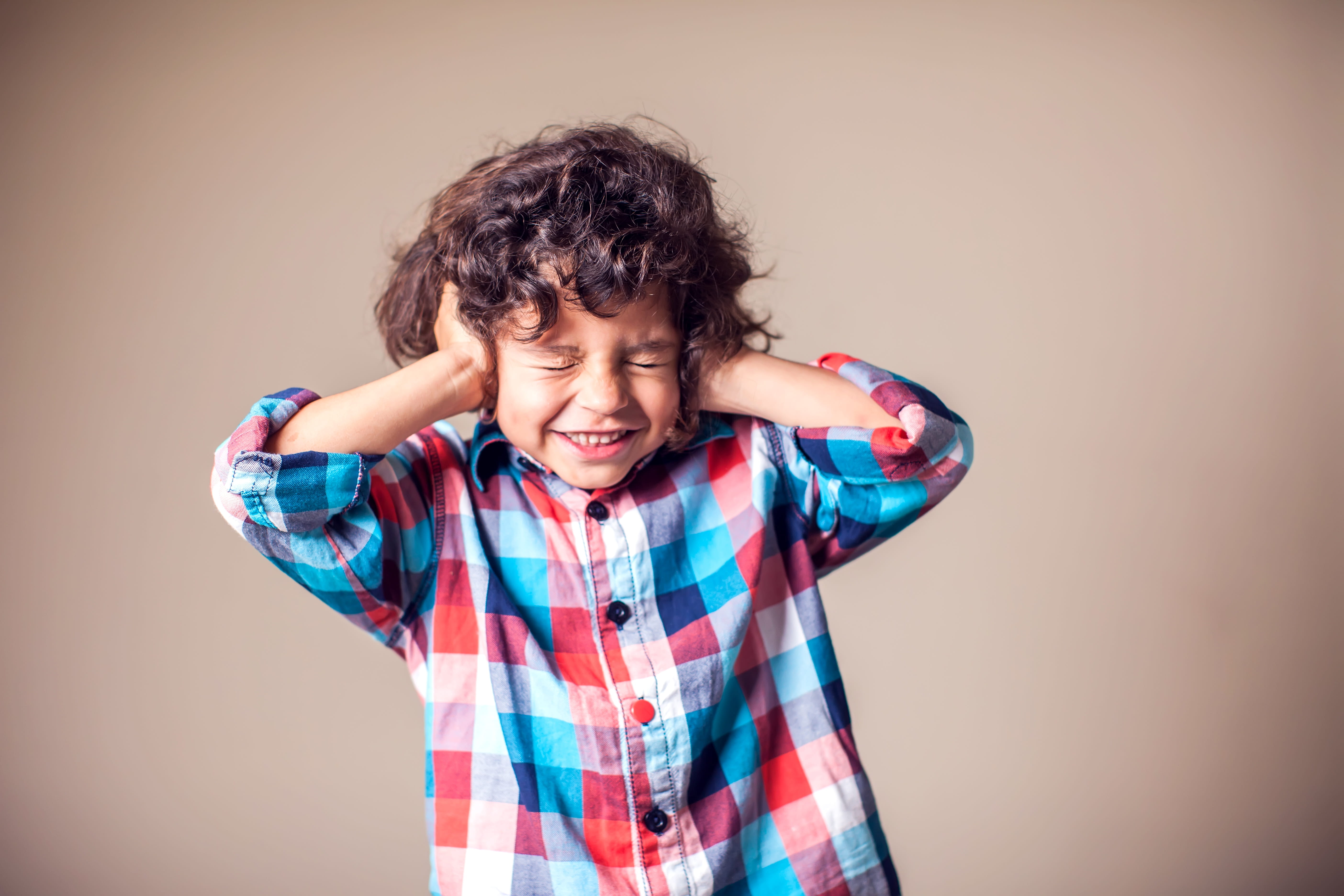 A young boy in a plaid shirt with curly brown hair covers his ears with his hands.