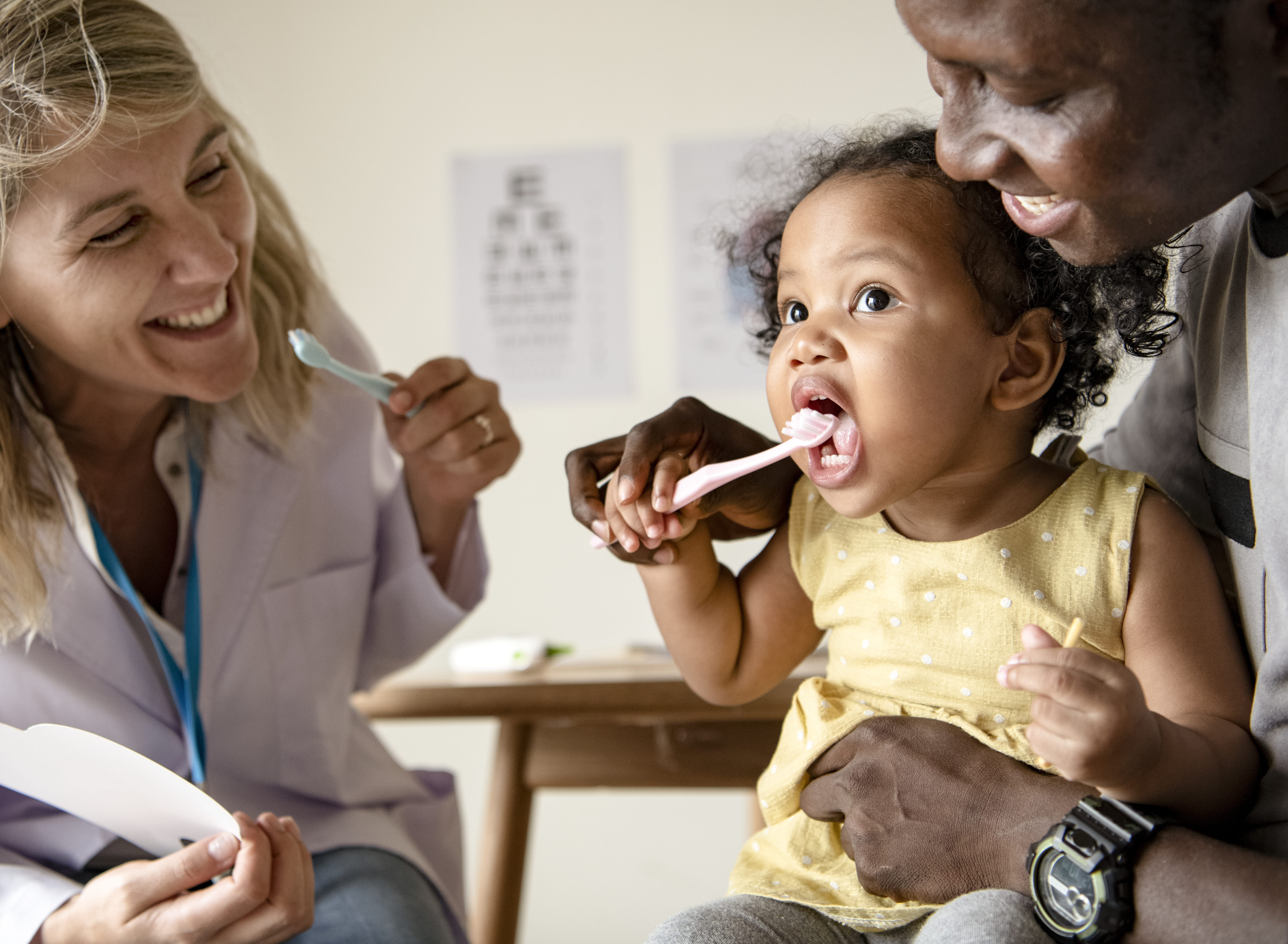 A young child practices brushing teeth with her parent and dentist assisting.