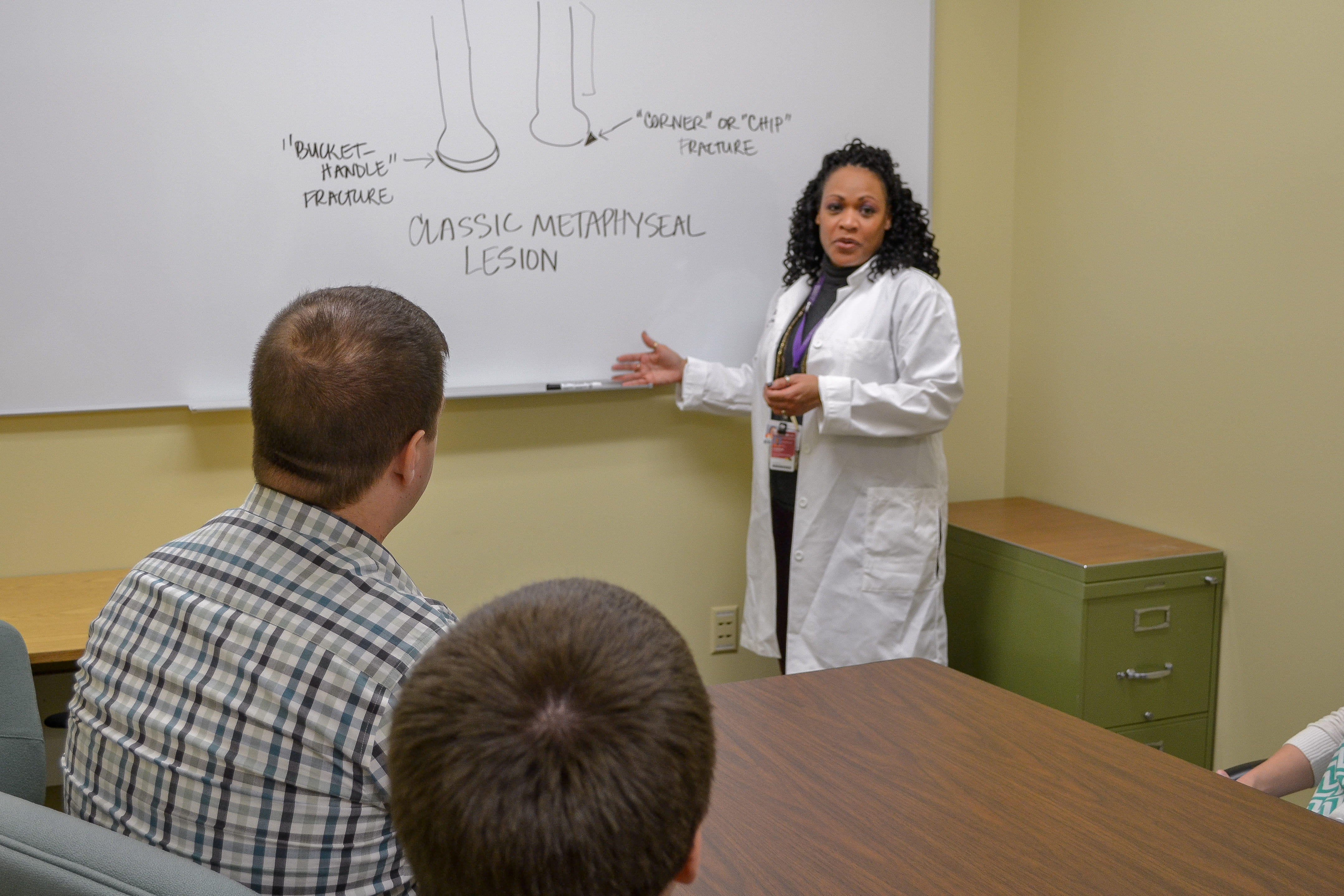 a faculty member in a white coat stands at a white board explaining something to a group of fellows