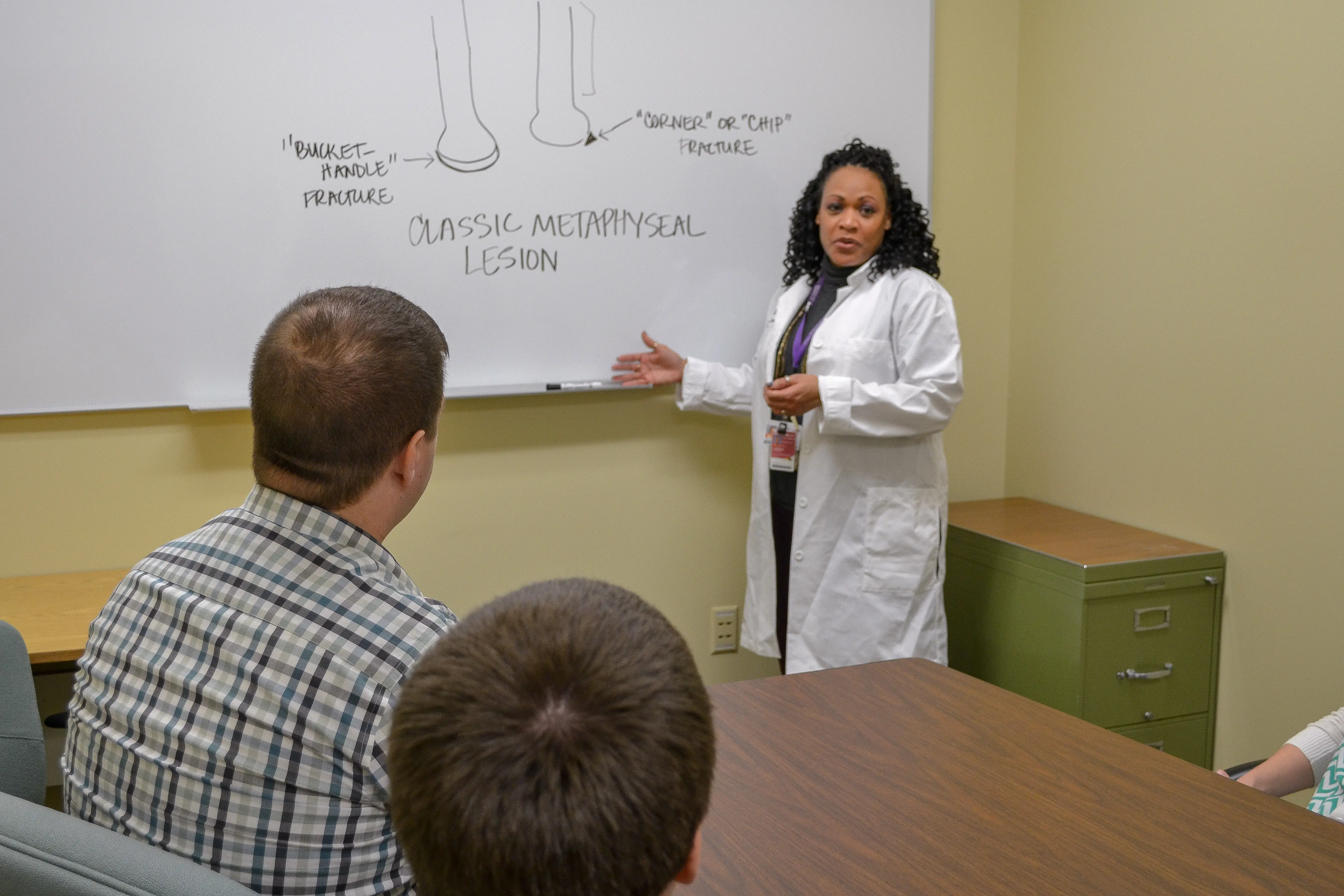 A woman in a white coat points to a hand drawn diagram on a white board at the front of a classroom.