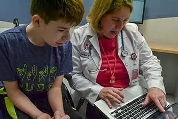 A physician wearing a white coat and stethoscope points to information on her laptop screen. A young patient sitting next to her is also looking at the screen.