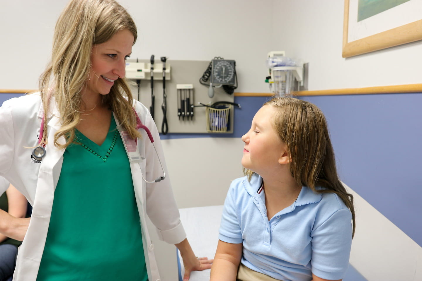 A physician with long blond hair speaks with a pediatric patient in the clinic. The doctor is wearing a white coat and green shirt. The patient has long light-brown hair and is wearing a blue shirt.
