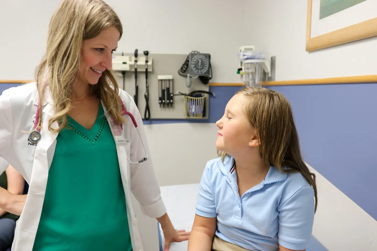 A physician with long blond hair speaks with a pediatric patient in the clinic. The doctor is wearing a white coat and green shirt. The patient has long light-brown hair and is wearing a blue shirt. 