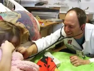 A physician smiles as he bends low to examine a young patient holding a teddy bear