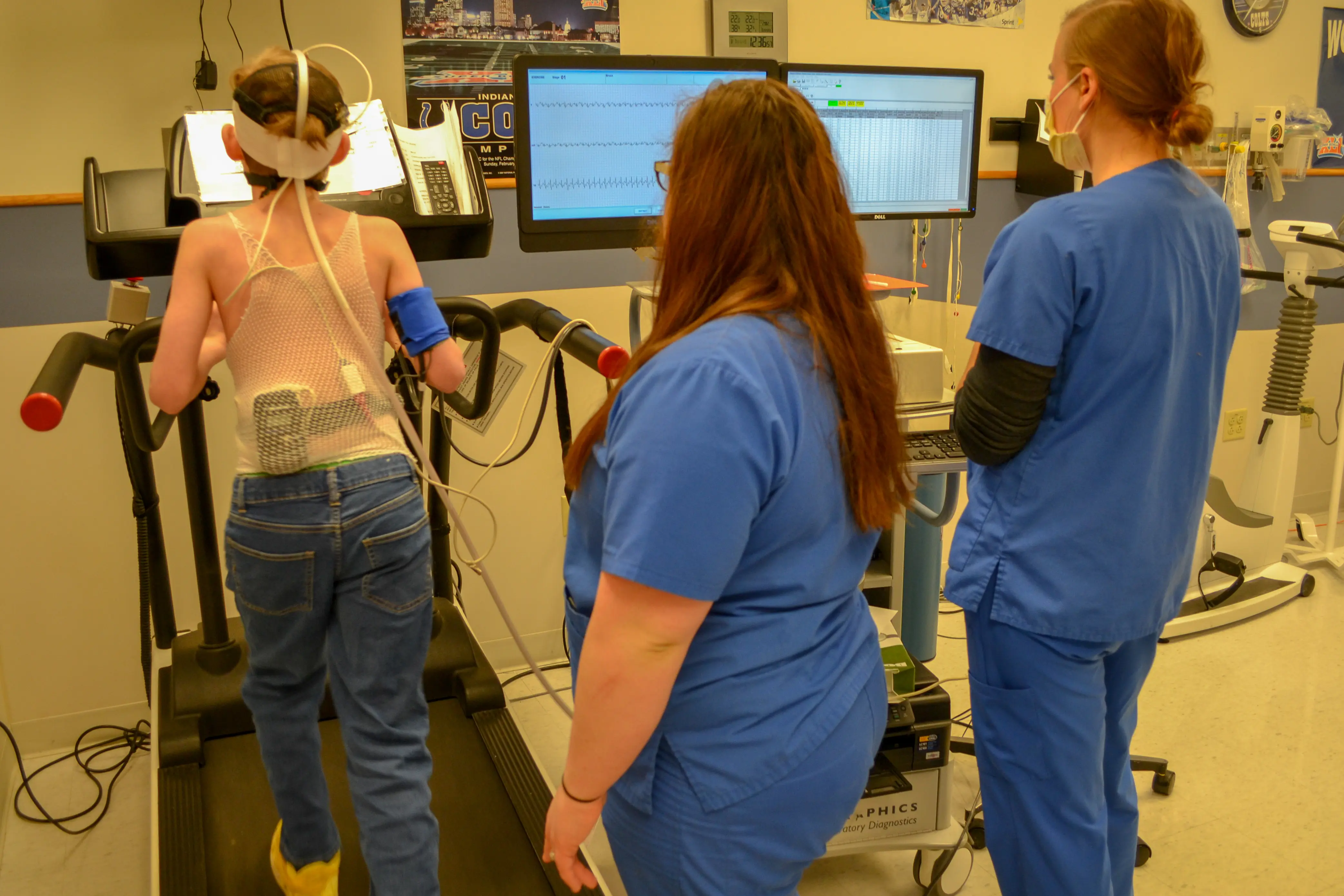 A young patient walks on a treadmill with two nurses standing by. He has monitoring equipment on his head and torso.