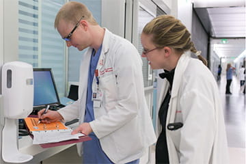 Two doctors standing near computer in hospital hallway reviewing paperwork