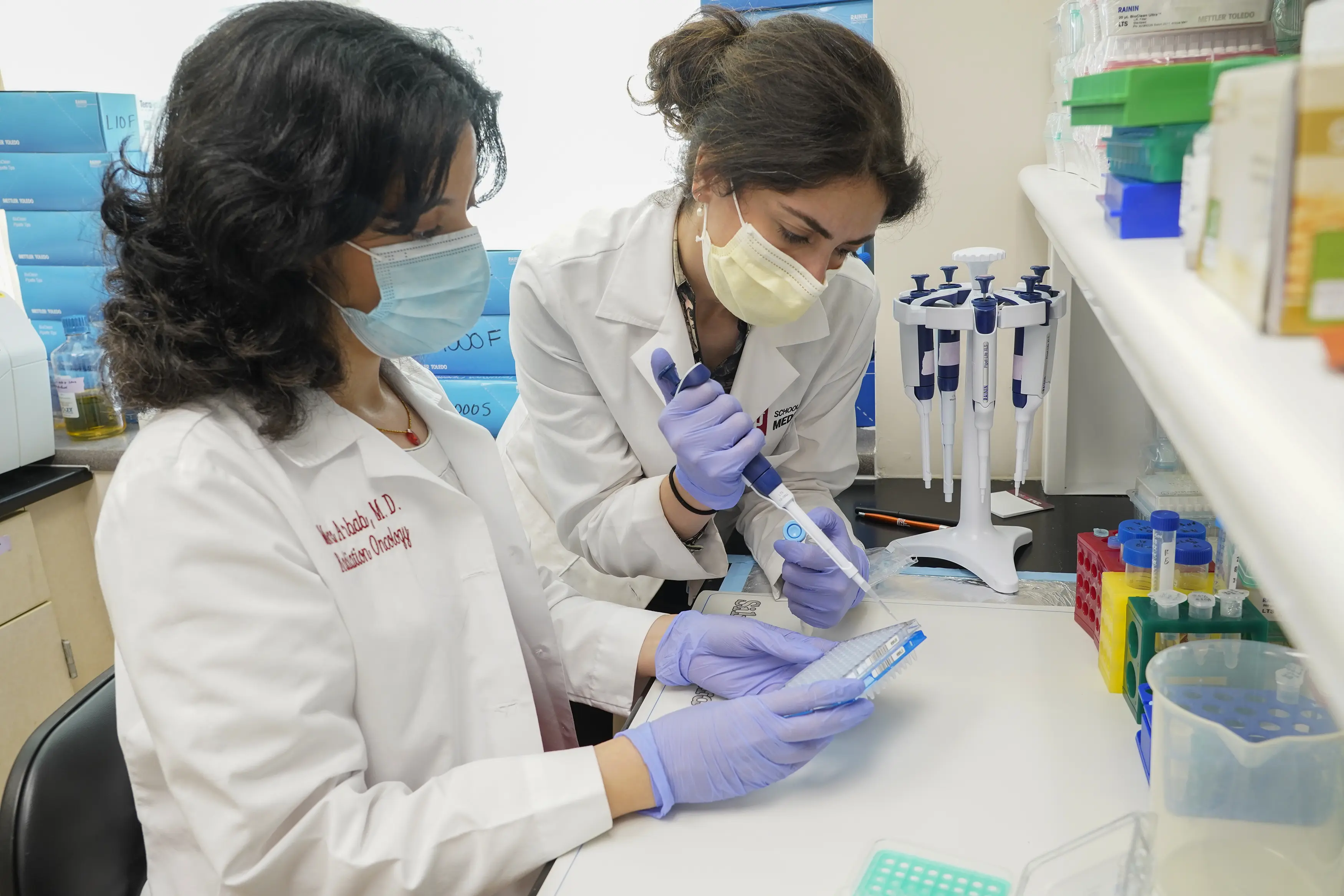 Two researchers in masks, lab coats and gloves work together to prepare lab samples