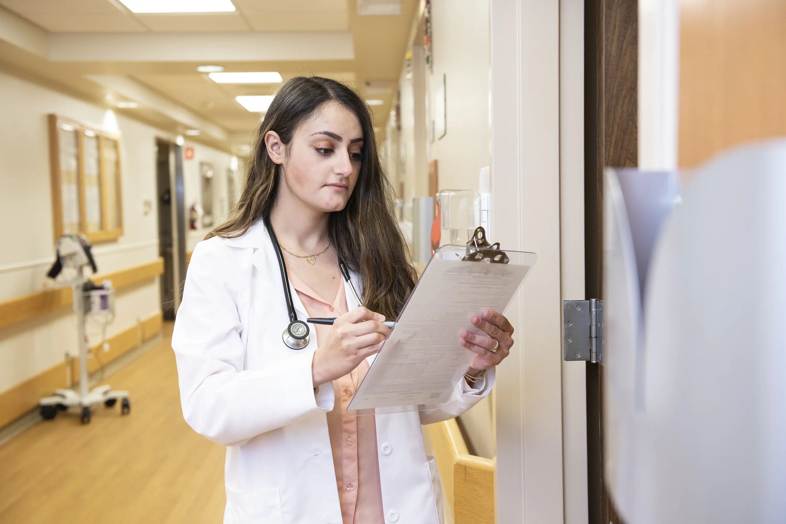 A woman wearing a white coat and stethoscope reviews patient information outside of a hospital room