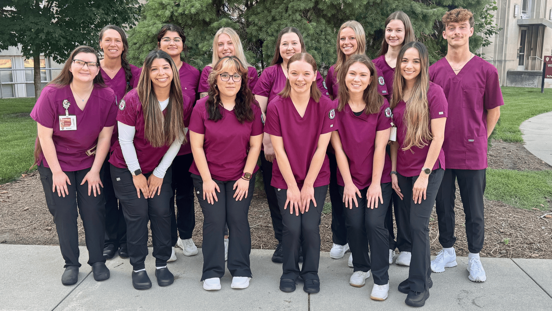  A group of 13 young adults, appearing to be students, are standing outside in front of some evergreen trees and a building. They are all wearing matching maroon scrub tops and dark pants. They are smiling and posing for the camera.