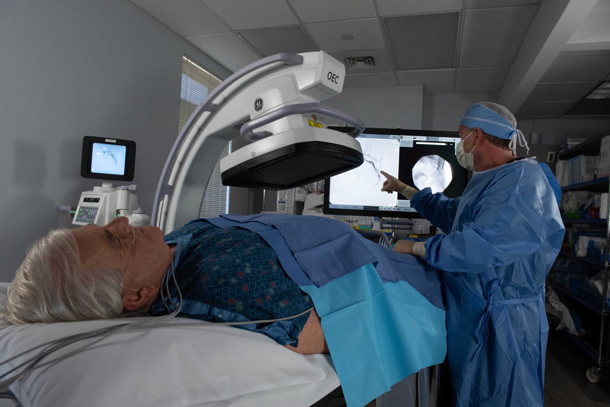 A medical professional in blue scrubs, a mask, and a surgical cap stands on the right, pointing at a large monitor displaying a medical image, possibly an X-ray. A patient with white hair lies on their back on a gurney, covered by blue drapes, with a nasal cannula visible. Above the patient, a large, curved GE OEC imaging machine is positioned. Another monitor is visible in the background to the left. The scene appears to be an interventional radiology suite or operating room.