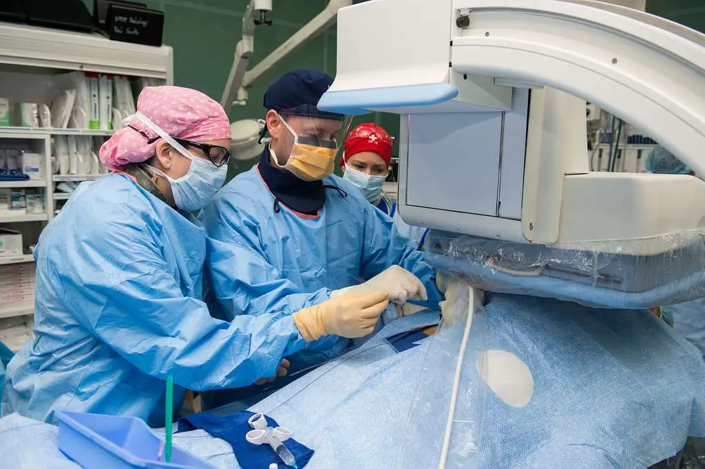 Two surgeons, wearing surgical gowns, masks, and caps, stand on either side of a draped patient, looking down at the surgical site. The surgeon on the left wears a pink surgical cap and glasses, while the surgeon on the right wears a blue surgical cap and a yellow mask. A third person, also in scrubs, a red cap, and a mask, is visible in the background, looking towards the patient. Directly over the patient, a large, white and blue medical imaging machine is positioned, with its screen facing the surgeons. The room appears to be an operating room, with medical supplies visible on shelves in the blurry background to the left.