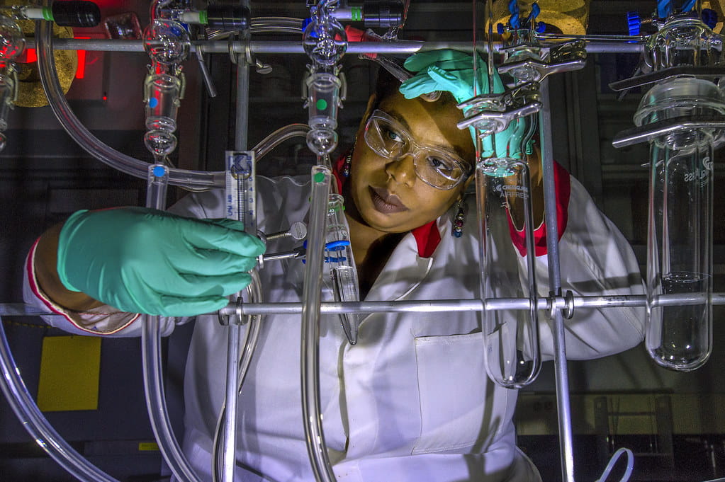  A female scientist wearing a white lab coat, green gloves, and clear safety glasses is meticulously working with complex laboratory glassware and tubing, with various liquids visible within the apparatus. She is looking intently at the setup, specifically at a syringe or pipette she is holding to make an adjustment. The background is dark, and the equipment is illuminated, highlighting the intricate scientific process.
