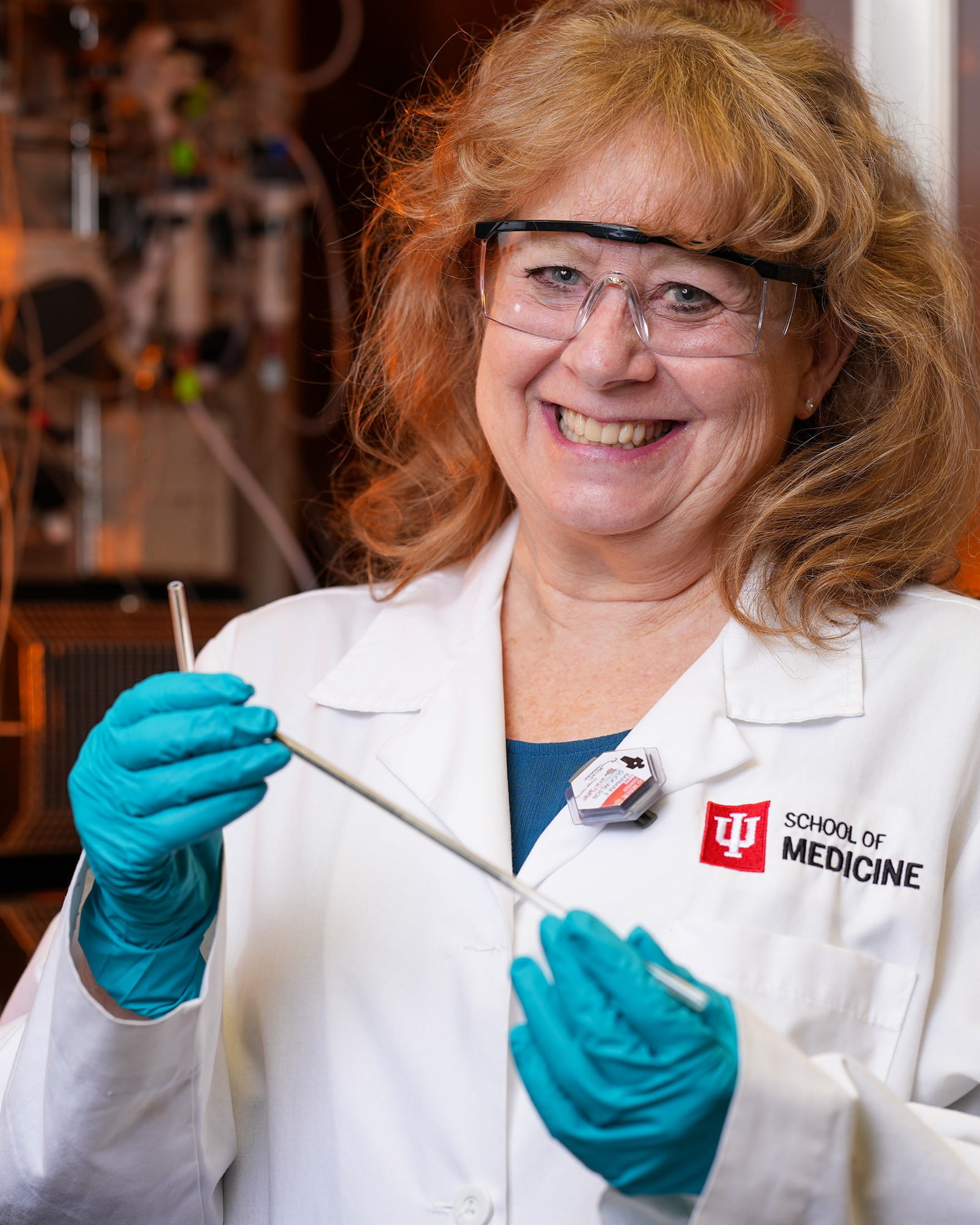 female scientist smiling in a white coat and lab glasses while holding a sample.