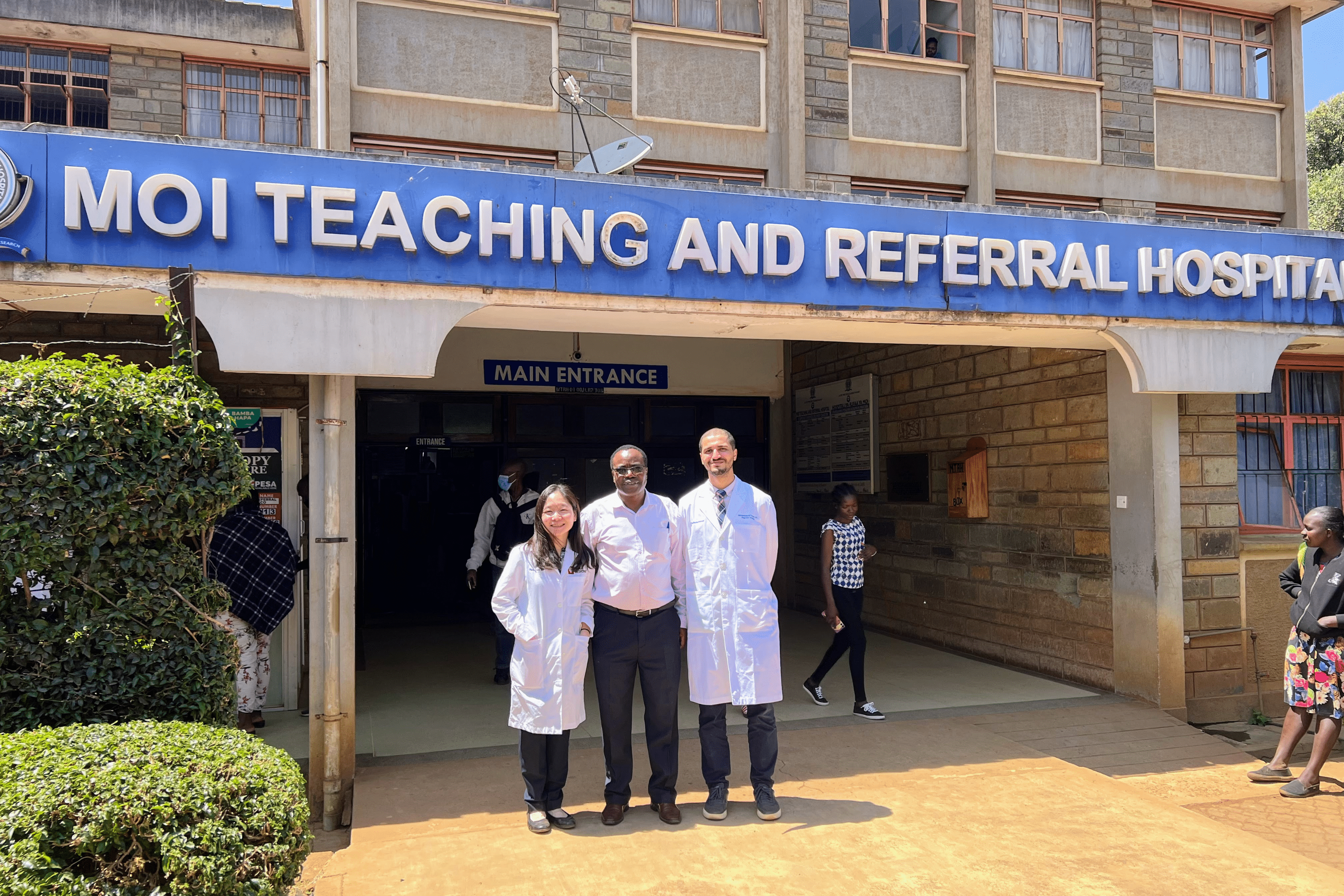  A wide-shot photo of two doctors, one woman and one man, posing with a third man in front of a building with a large blue sign that reads "MOI TEACHING AND REFERRAL HOSPITAL"