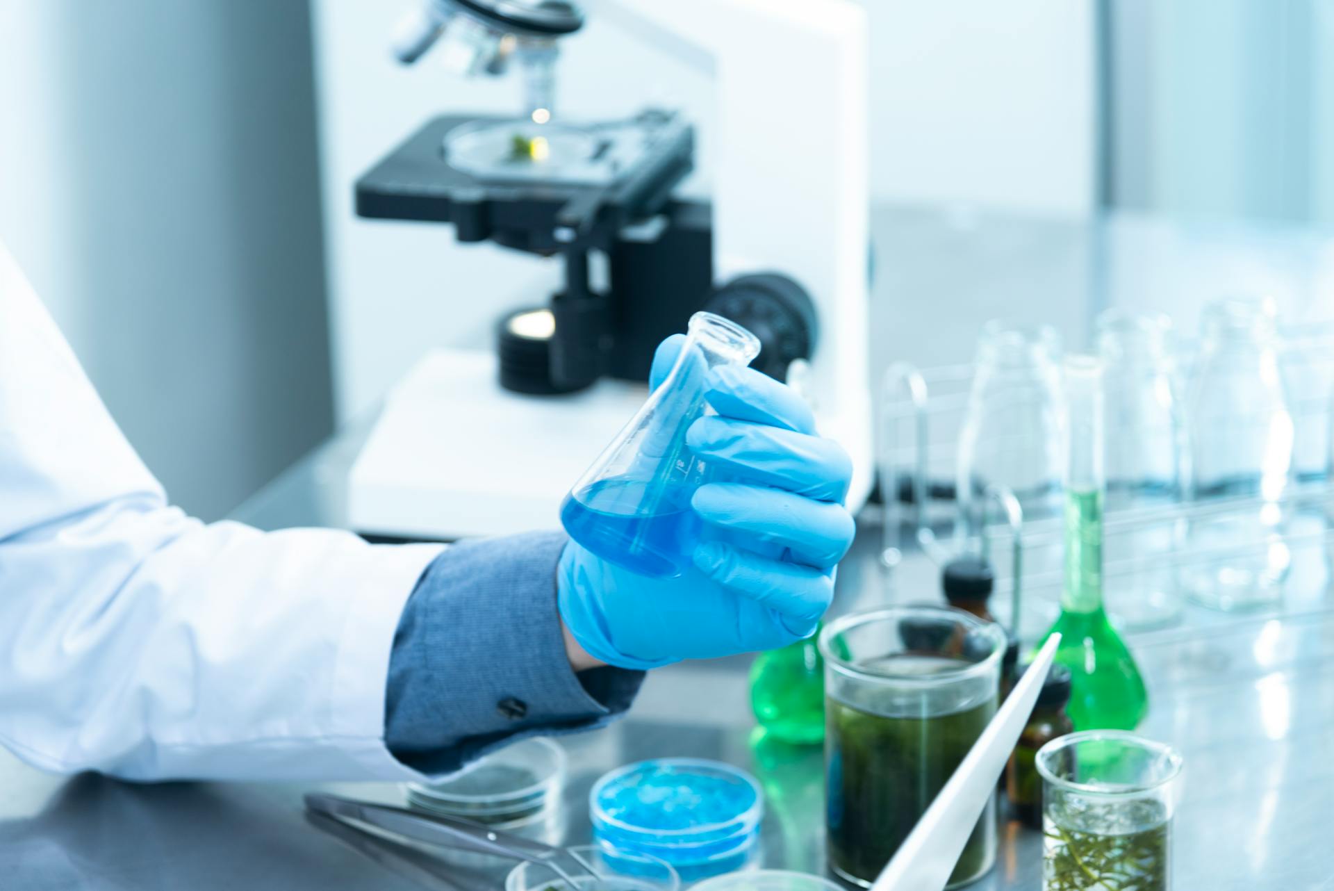 A blue-gloved hand hold a beaker with blue liquid. Other beakers are in the background, full of green liquid. There is also a microscope in the background.