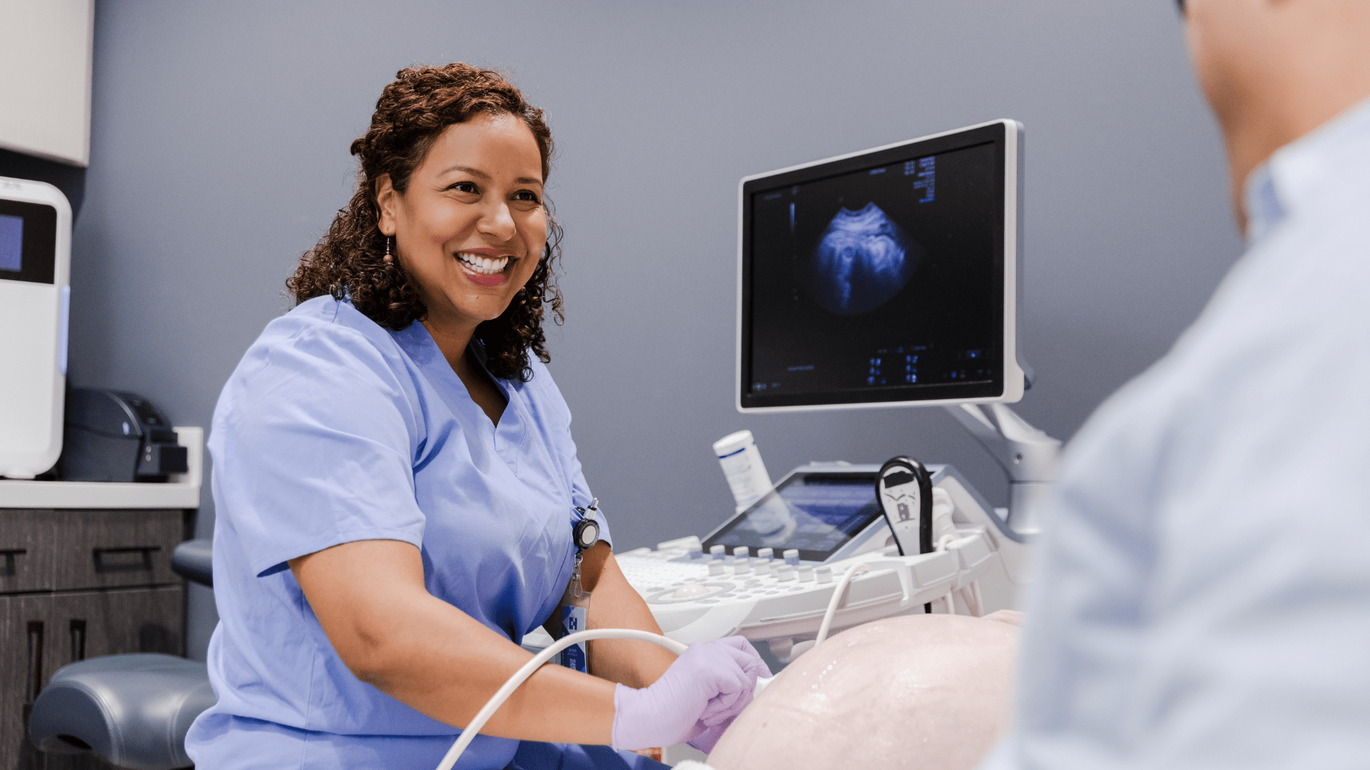 A smiling sonographer, wearing light blue scrubs and purple gloves, performs an ultrasound on a patient. A large monitor displaying an ultrasound image is visible in the background. 