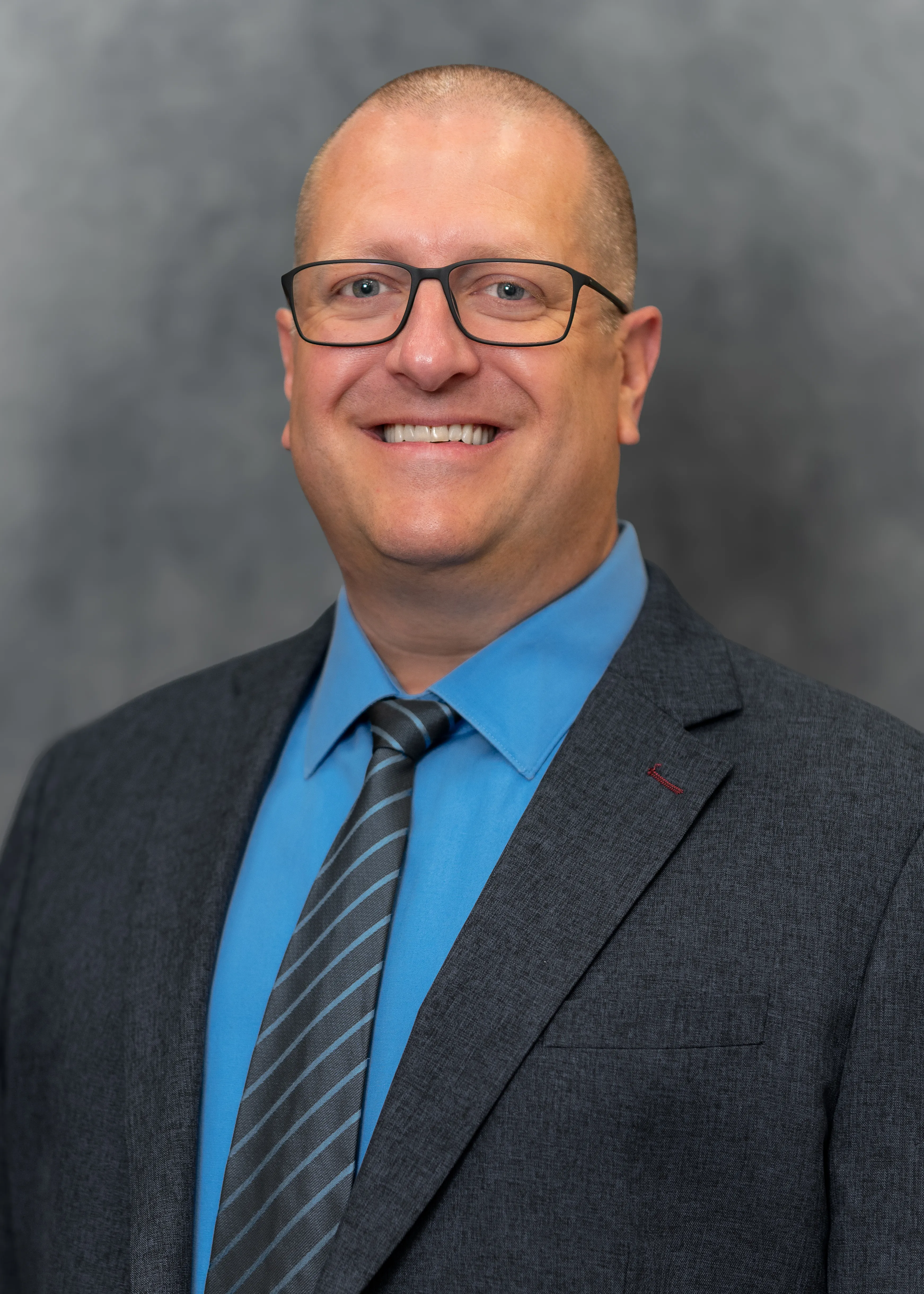 Professional headshot of a smiling man with closely cropped hair and black-framed glasses, wearing a dark gray suit, blue shirt and striped tie against a neutral gray background.