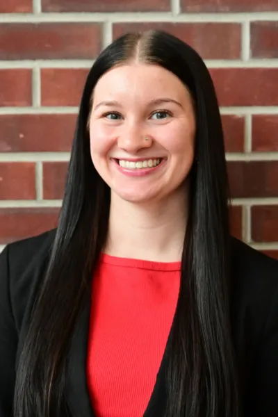 Professional heashot of Caitlin Burns on a brick wall background dress in a red top and black blazer