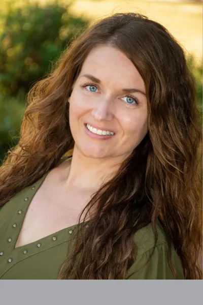 Professional outdoor portrait of a smiling woman with long, wavy brown hair, wearing an olive green top against a softly blurred natural background.