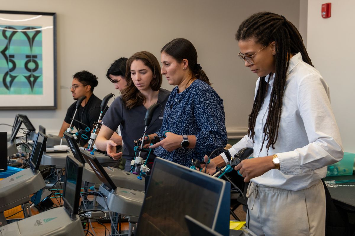 Payton Miller, MD, points to a surgical simulation console while standing with four other trainees in a clinical skills lab. The group stands side by side at a row of laparoscopic training stations with monitors and handheld instruments, focusing on the equipment as they practice.