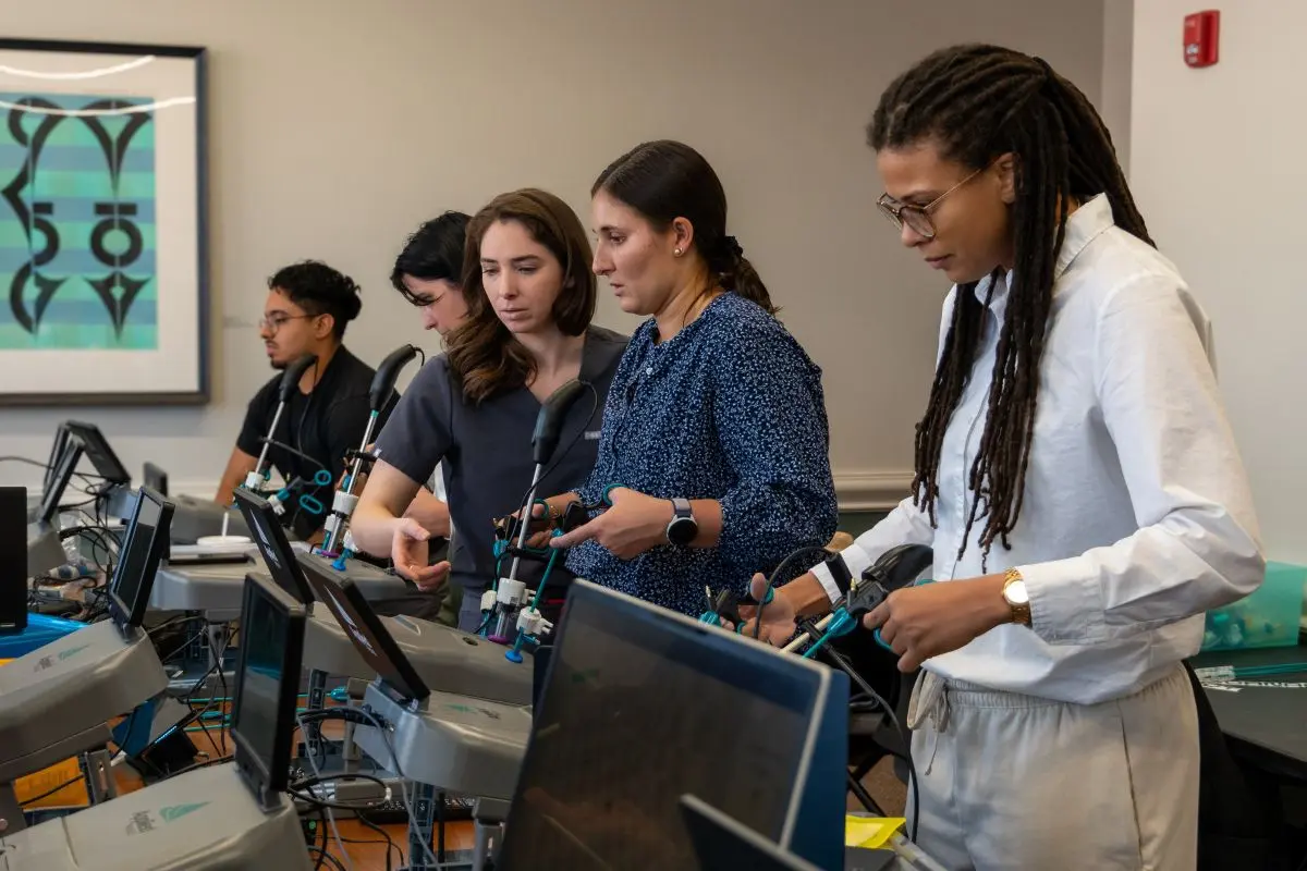 Payton Miller, MD, points to a surgical simulation console while standing with four other trainees in a clinical skills lab. The group stands side by side at a row of laparoscopic training stations with monitors and handheld instruments, focusing on the equipment as they practice.