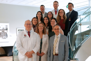 Thirteen medical professionals and trainees pose together on an indoor staircase in a bright, modern building. Three people in the front row stand closest to the camera, including two wearing white lab coats and one in a gray suit with a yellow tie. Others stand on the steps behind them, some in lab coats and others in business attire. All are facing forward and smiling.