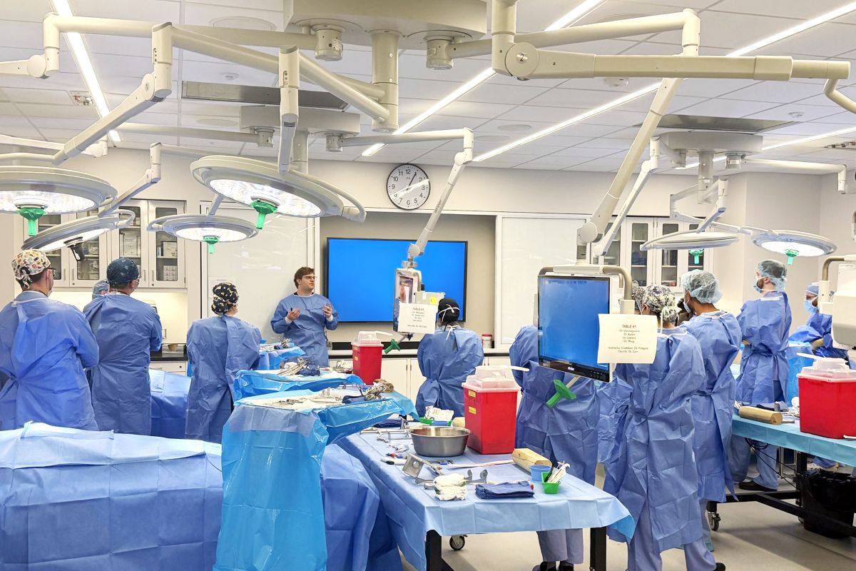Dr. Lukowski stands at the front of a surgical skills laboratory leading the annual ASSET course. PGY-3 general surgery residents, dressed in surgical gowns, caps, and masks, are gathered around medical table stations equipped with surgical instruments, monitors, and overhead operating lights.