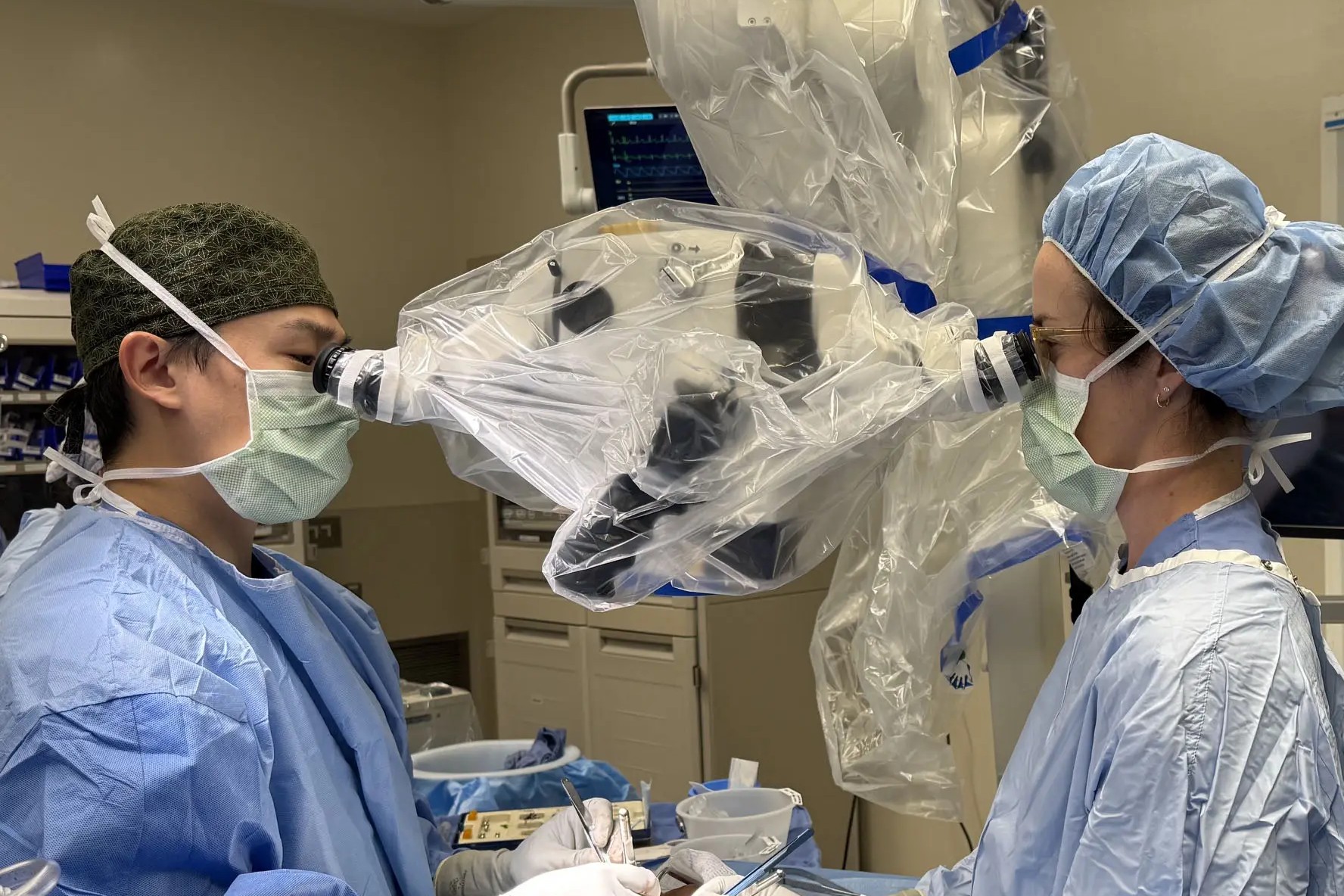 One male and one female surgeon look through an enhanced loupe to perform a microsurgery on a paitnet. 
