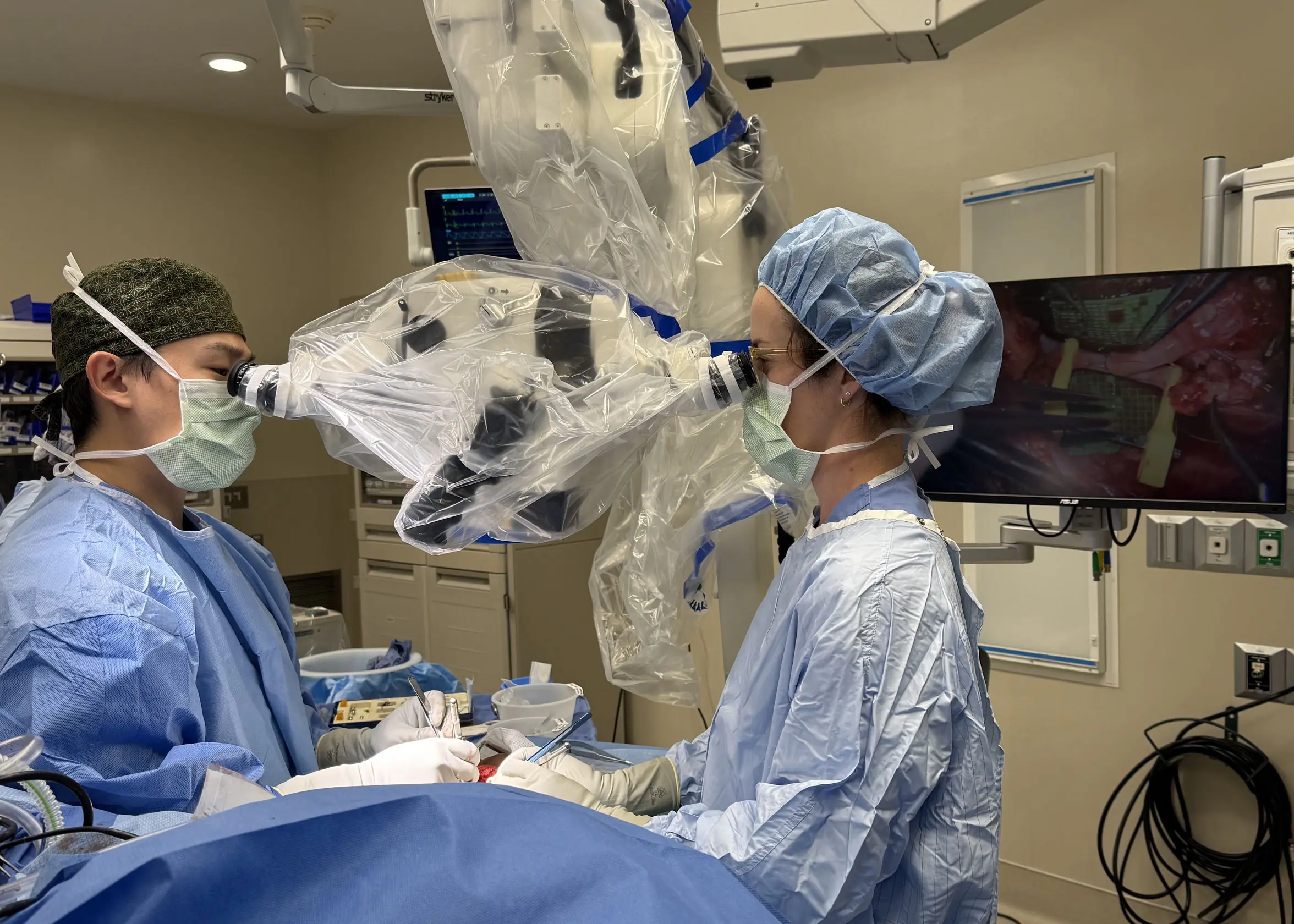 One male and one female surgeon look through an enhanced loupe to perform a microsurgery on a paitnet. 