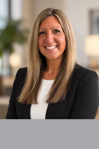 Professional portrait of a smiling woman with long, straight blonde hair, wearing a black blazer over a white top against a softly blurred indoor background.