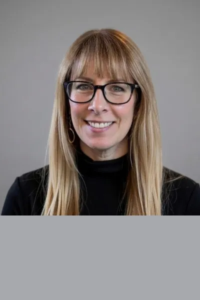 Professional headshot of a smiling woman with long blonde hair and bangs, wearing black-framed glasses and a black top against a neutral gray background.