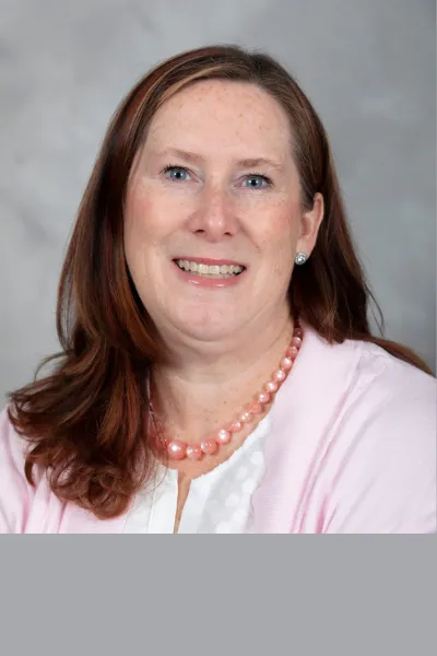 Professional headshot of a smiling woman with long auburn hair, wearing a light pink cardigan, white top and pink beaded necklace against a neutral gray background.