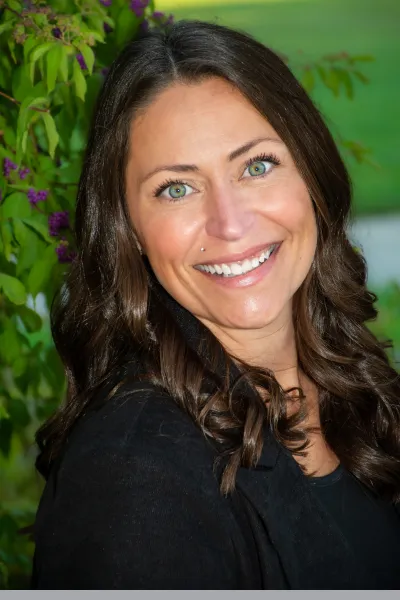 Professional portrait of a smiling woman with long, wavy dark brown hair, wearing a black top against a softly blurred green, leafy background