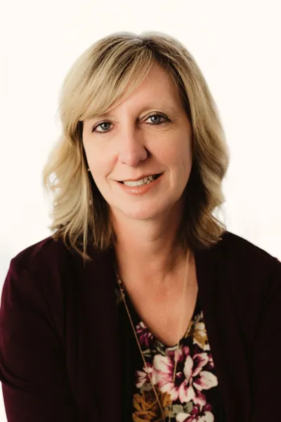 Professional headshot of Michelle Artmeier smiling, wearing a dark blazer and floral shirt on a solid warm white backdrop