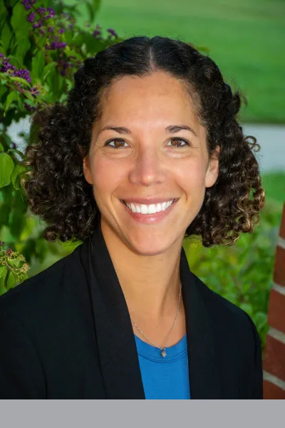 Professional portrait of a smiling woman with short, curly dark hair, wearing a black blazer over a blue top against a softly blurred green, leafy background.