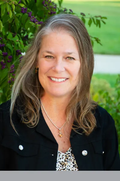 Professional outdoor portrait of a smiling woman with long gray-brown hair, wearing a black jacket and pendant necklace against a leafy green background.