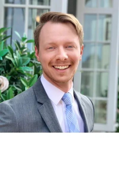 Professional portrait of a smiling man with short light brown hair and a trimmed beard, wearing a gray suit and light blue tie in a bright indoor setting with greenery.