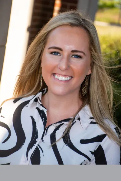 Professional outdoor portrait of a smiling woman with long blonde hair, wearing a black-and-white patterned blouse and dangling earrings.