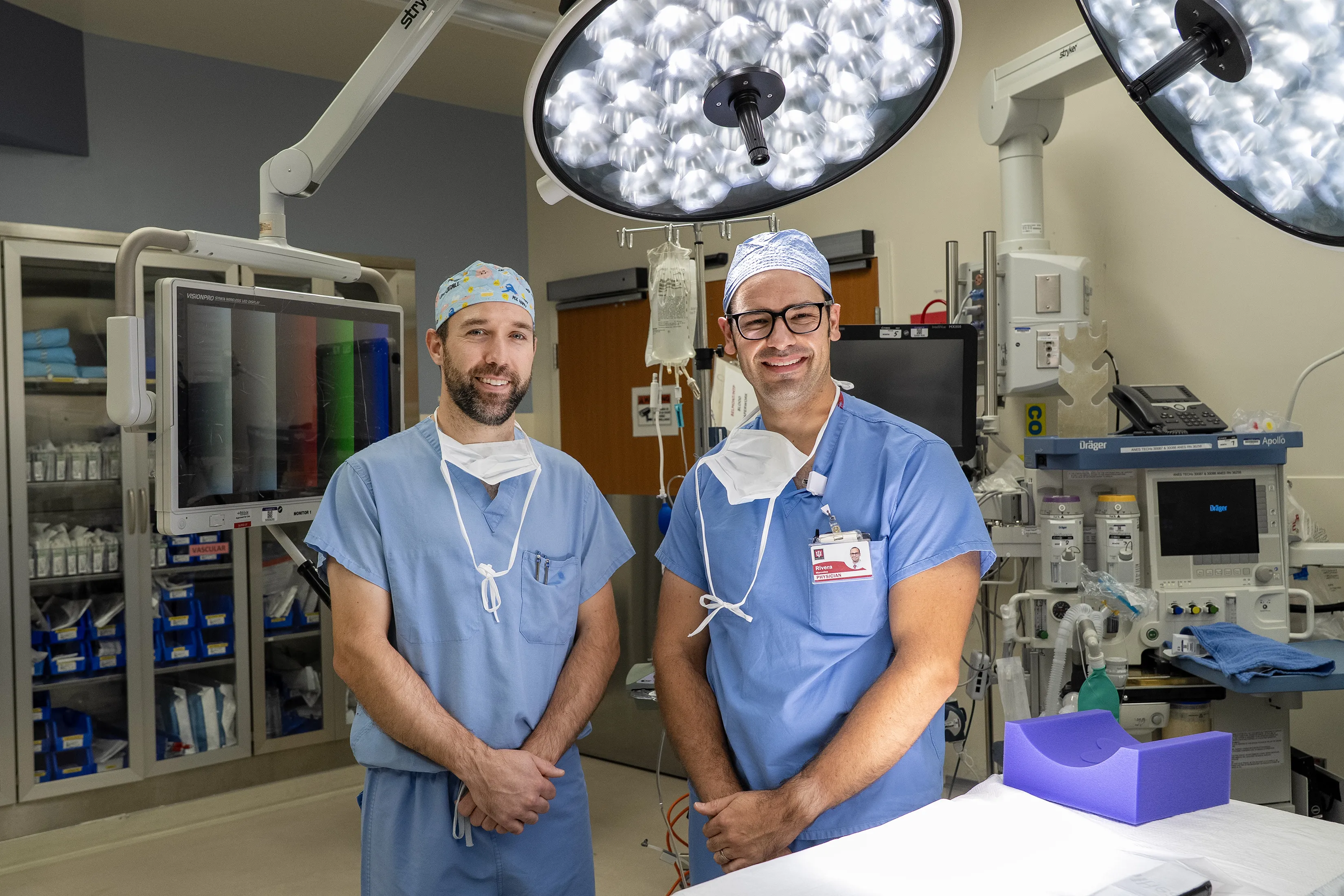 Dr. Thomas Shelton and Dr. Marcelino Rivera stand together in an operating room wearing blue surgical scrubs and caps.