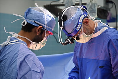 two surgeons wearing protective scrubs, goggles, masks and hair covering work together in the operating room.