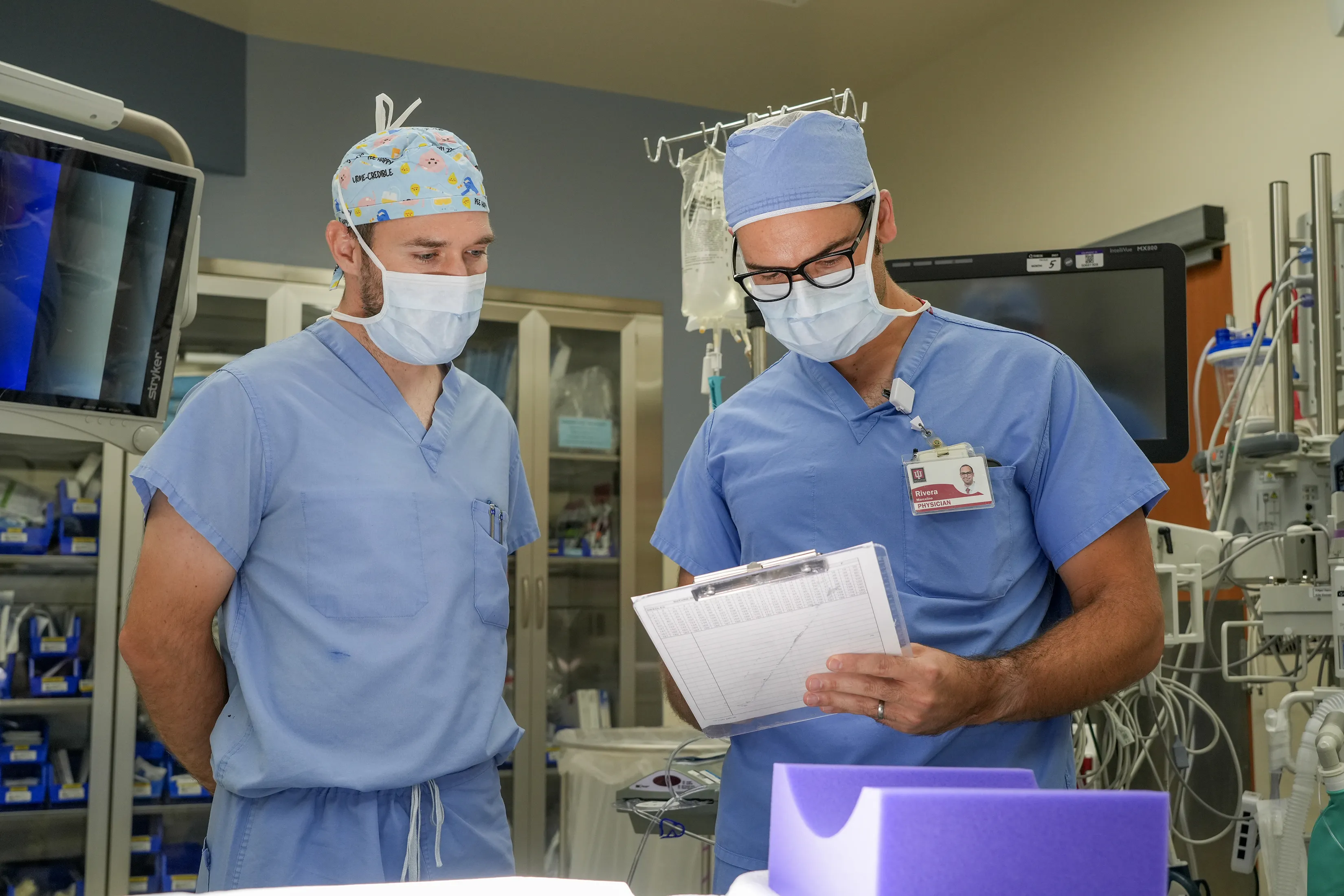 Two medical professionals in surgical scrubs and masks review a clipboard together in an operating room.