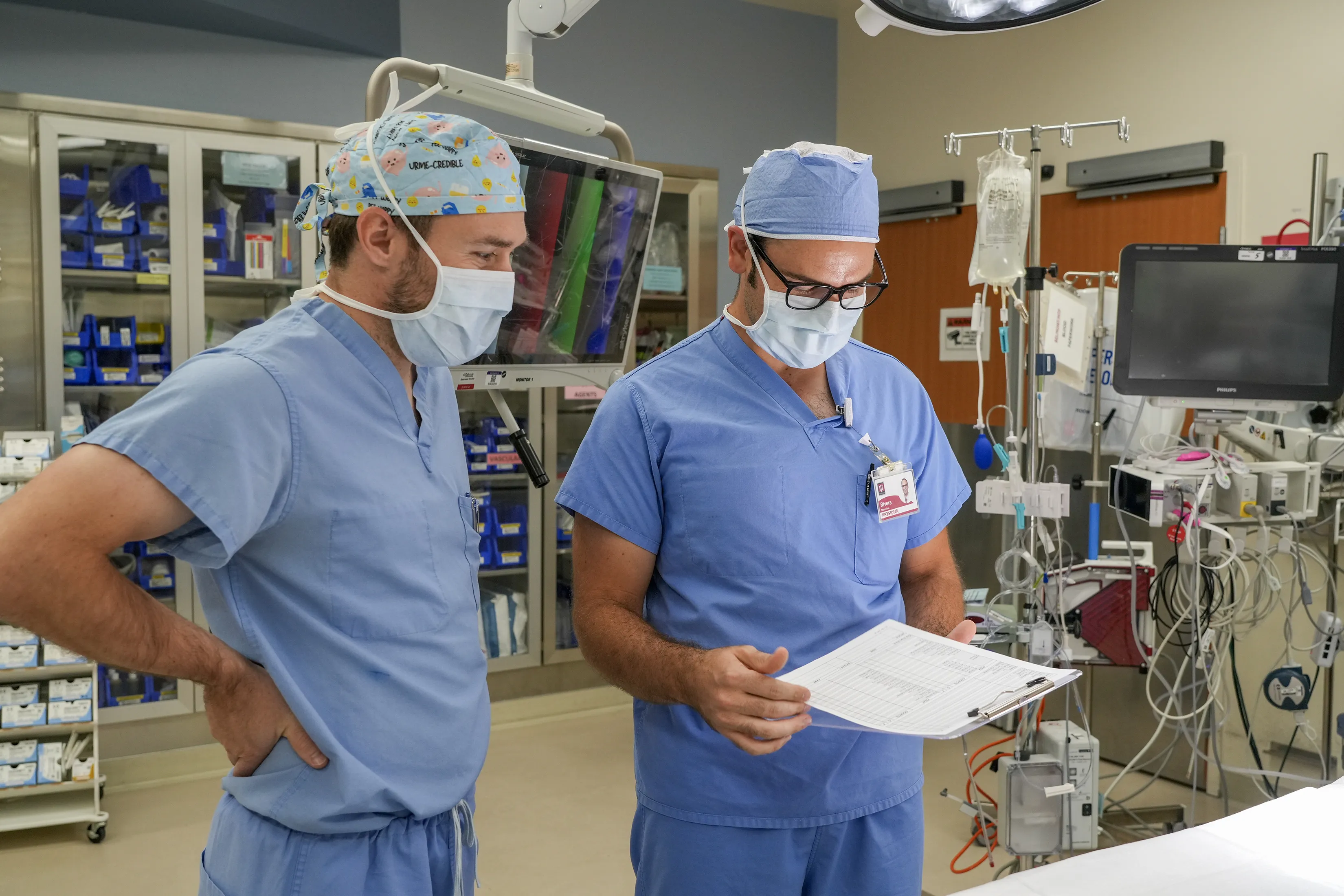 Two medical professionals in surgical scrubs and masks review a clipboard together in an operating room.
