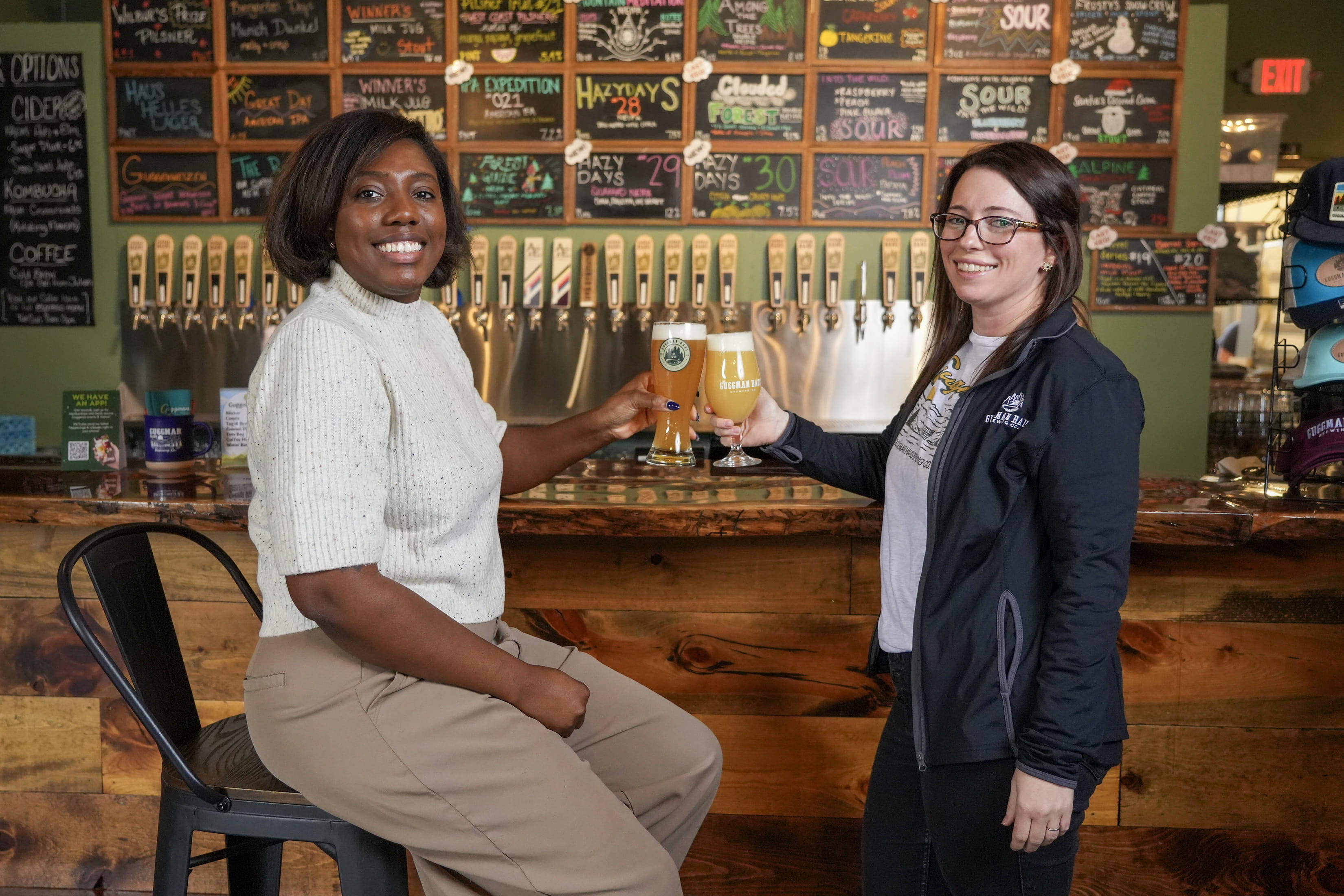 Sacha Sharp and Abby Gorman stand at the bar in Guggman Haus Brewery. Each woman is holding a glass of beer and smiling. 